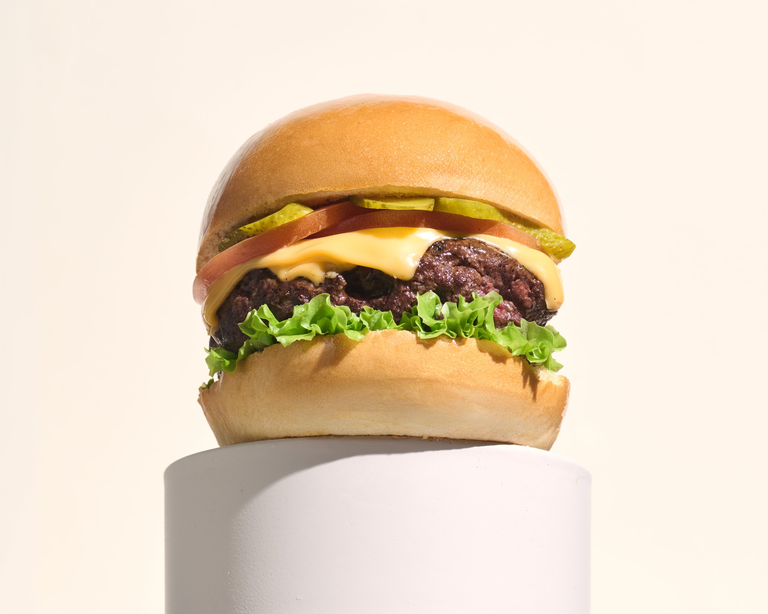 Hamburguesa Lusitana con queso, tomate y pepinillos sobre pedestal blanco